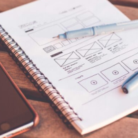 Gros plan sur un carnet contenant des croquis de wireframes, accompagné d’un smartphone posé sur un bureau en bois, illustrant le travail de design UX/UI.